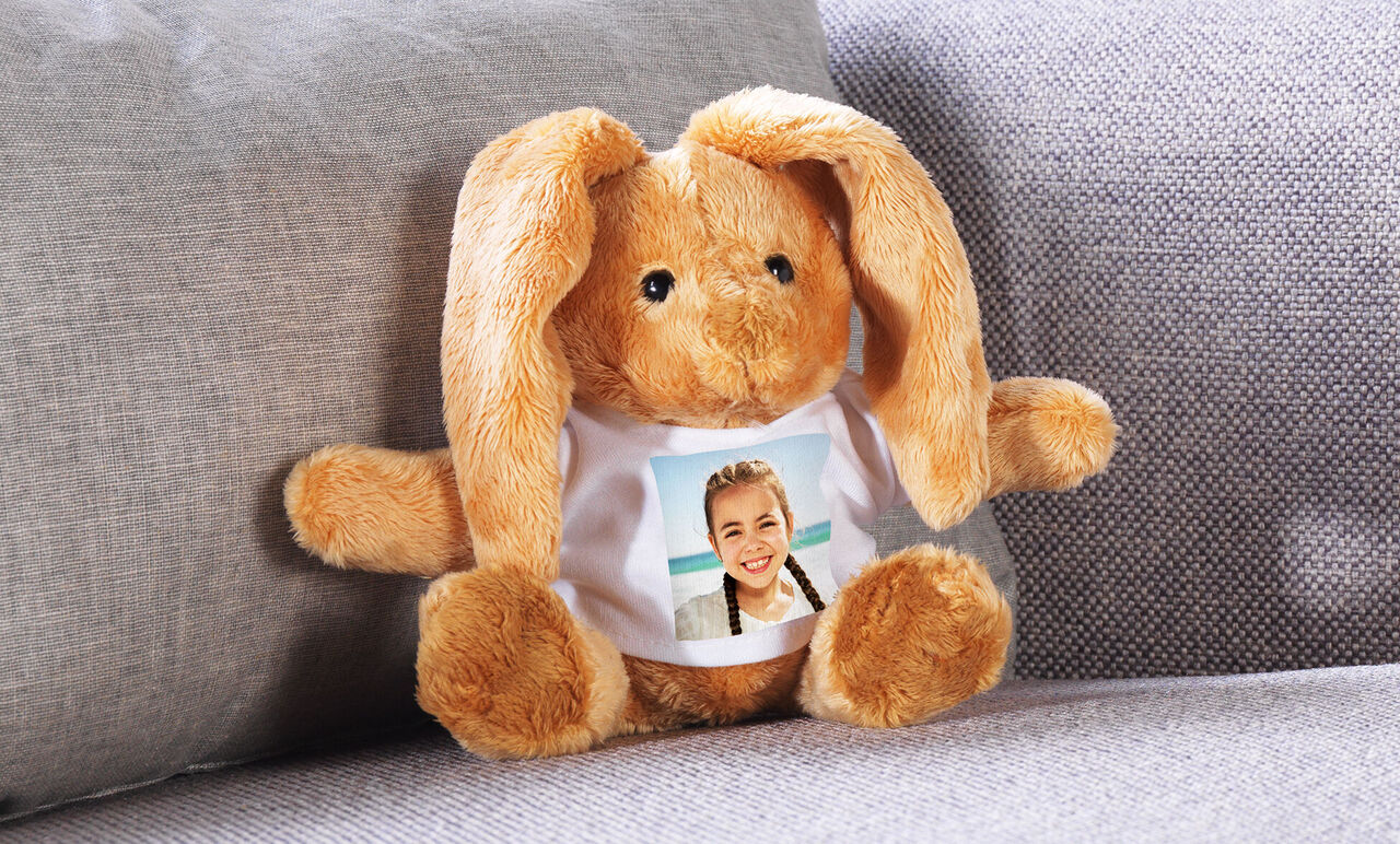 Un adorable lapin en peluche est assis sur un canapé. Il porte un T-shirt avec une photo.