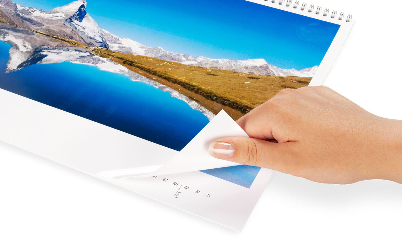 Une main feuillette un calendrier photo CEWE avec une belle vue de montagne.
