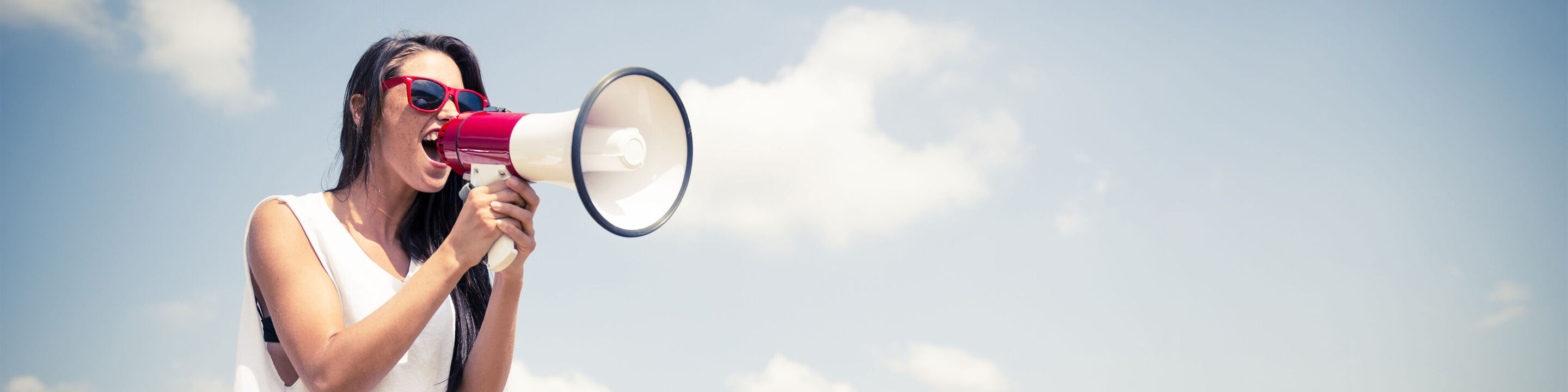 Une personne avec un mégaphone pose sous un ciel bleu avec quelques nuages.