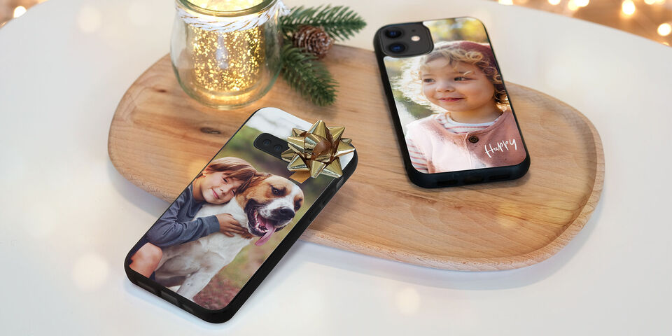 Deux coques de smartphone avec des designs photographiques sur une planche en bois. Une coque montre un chien avec un enfant et l'autre une mise en page personnelle pour une personne spéciale. Vase avec lumières en arrière-plan.