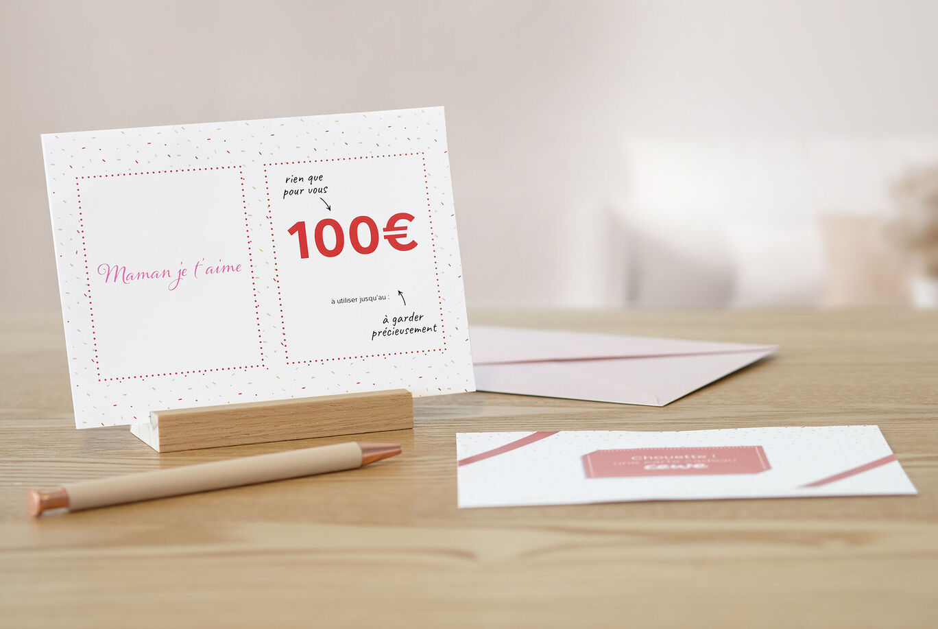 Une carte de vœux CEWE dans un étui-cadeau d'une valeur de 100 euros, sur une table avec un support en bois élégant et un crayon rose.