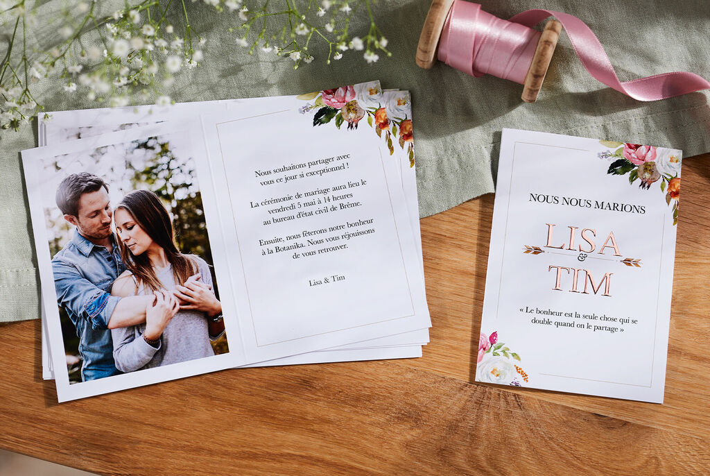 Un couple s'enlace dans la nature. En avant-plan, des invitations de mariage ornées de motifs floraux.