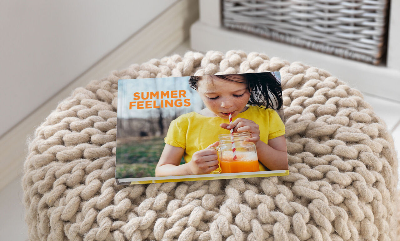 Une fille tient un verre avec une boisson colorée. En arrière-plan, un CEWE FOTOBUCH intitulé "Summer Feelings" sur un pouf confortable.