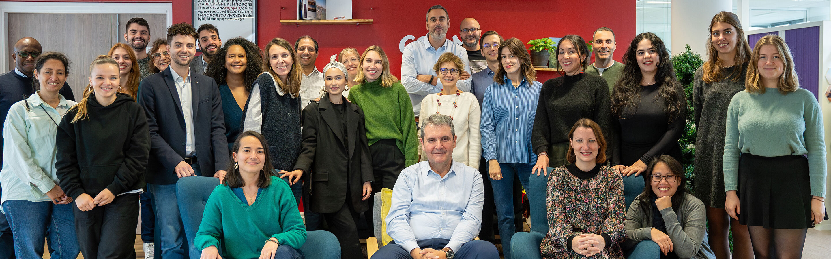 Photo d'équipe avec 27 personnes souriantes réunies. Environnement coloré.