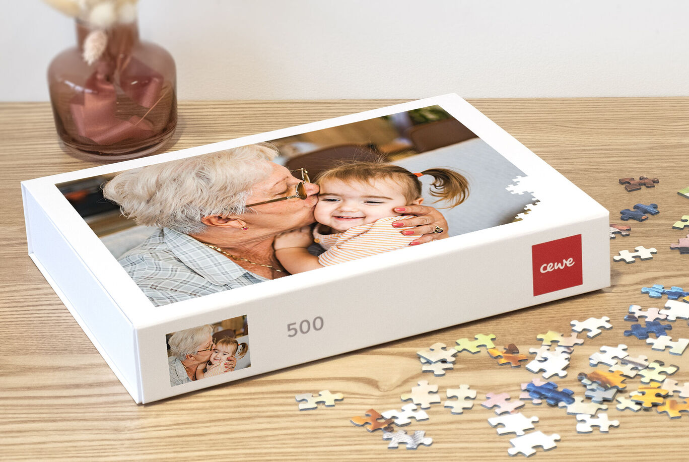 Une grand-mère souriante embrasse son petit-enfant joyeusement sur la joue. Une boîte de puzzle CEWE et des pièces de puzzle sont sur la table.