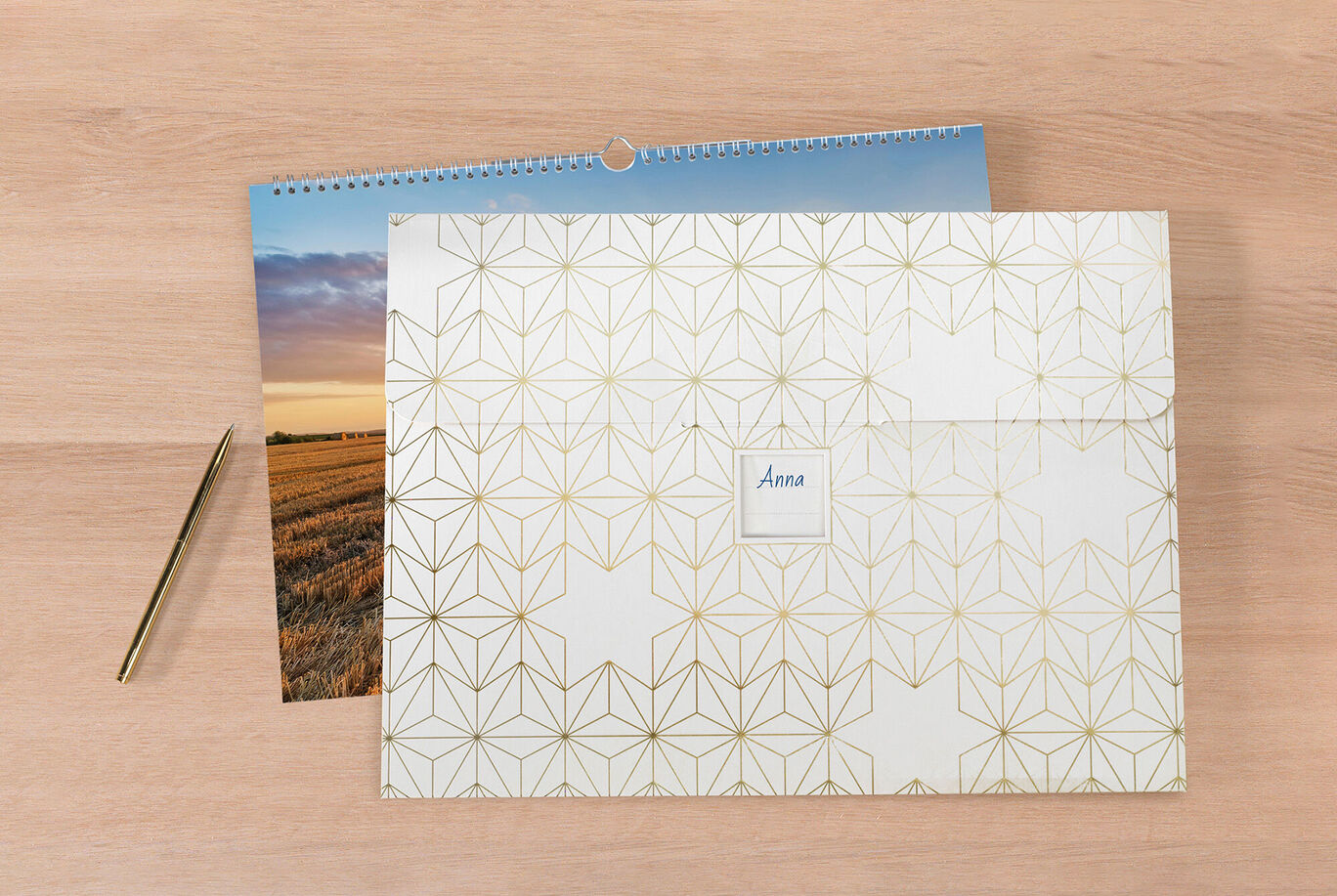 Un calendrier photo CEWE soigneusement conçu sur une table en bois, à côté une enveloppe décorative avec le nom 'Anna', et un stylo doré.