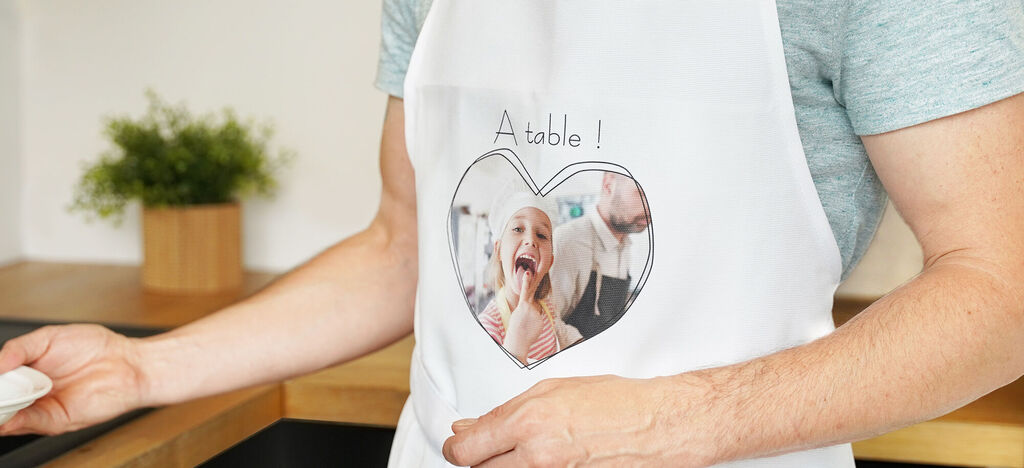 Un homme avec un tablier se tient dans la cuisine tenant un petit bol devant lui. L'apron montre une image en forme de cœur.