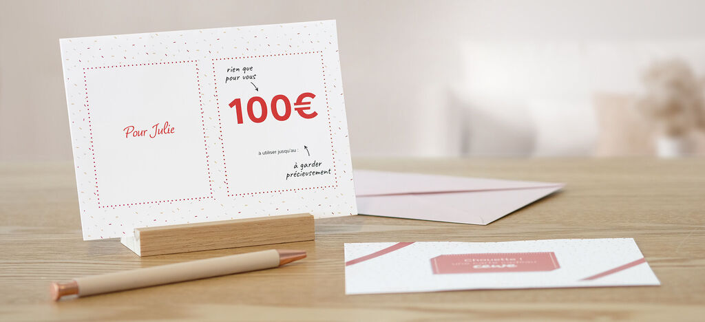 Cartes de vœux CEWE avec un bon de 100€ sur une table en bois, avec un stylo et une autre carte.
