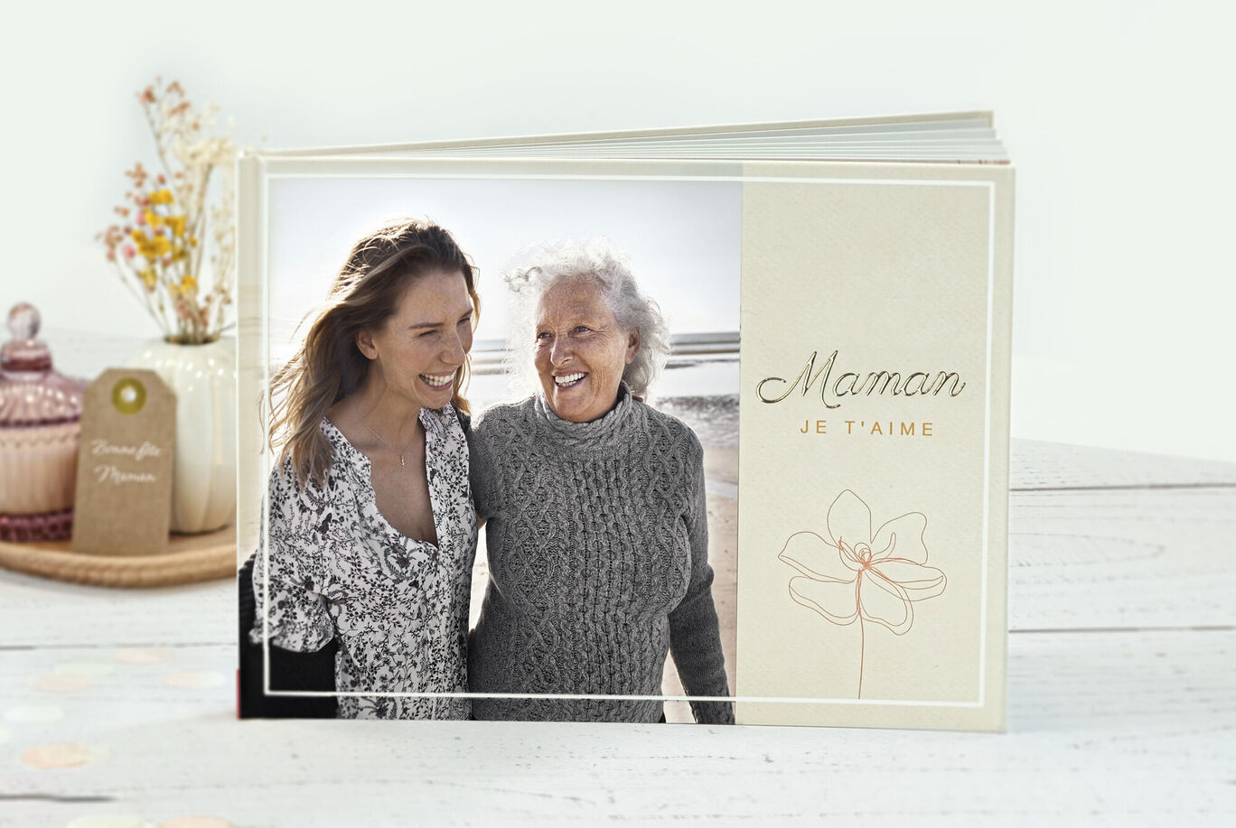 Deux femmes se tiennent sur la plage, s'enlaçant et souriant heureusement, à côté d'un album photo CEWE.