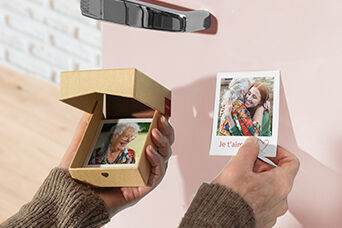 Une personne tient un petit cadeau avec une photo et un cœur. En arrière-plan, un élément décoratif et un mur.