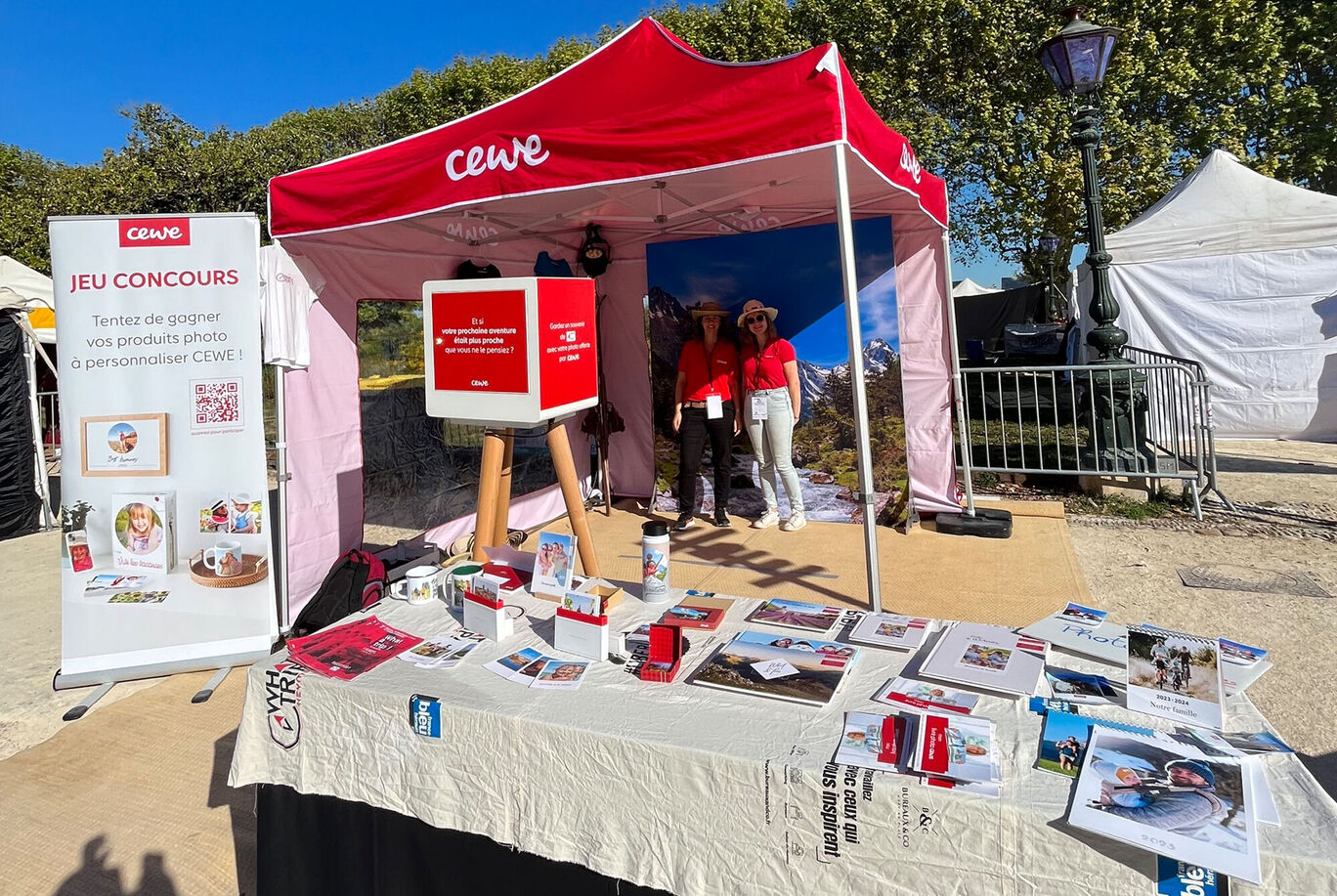 Stand CEWE avec panneau de compétition et plusieurs prospectus sur des produits photo.
