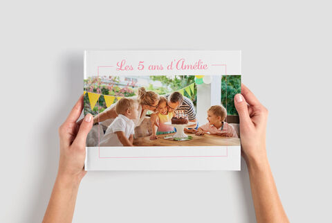 Des mains tiennent un album photo CEWE intitulé "Les 5 ans d'Amélie", et trois enfants regardent.