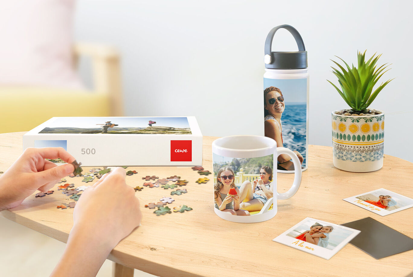 Une personne assemble un puzzle CEWE. Un gobelet et une bouteille avec des photos sont sur la table.