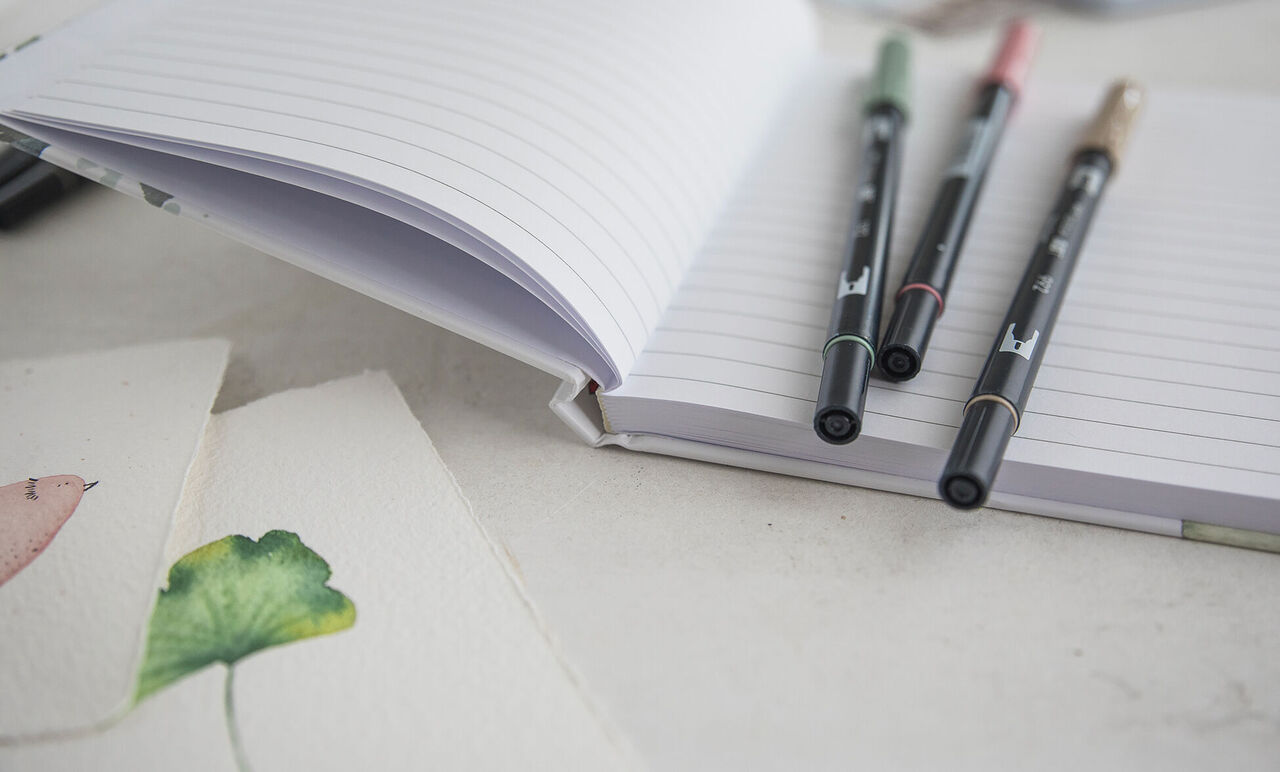 Cahier rigide ouvert avec trois stylos fins posés sur des pages lignées et illustrations sur des feuilles, ambiance créative.
