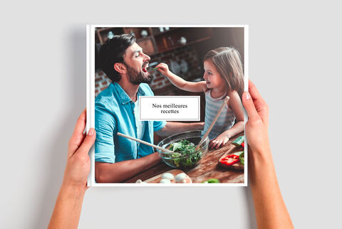 Une personne tient un livre photo CEWE avec des recettes. En arrière-plan, des ingrédients sont visibles.