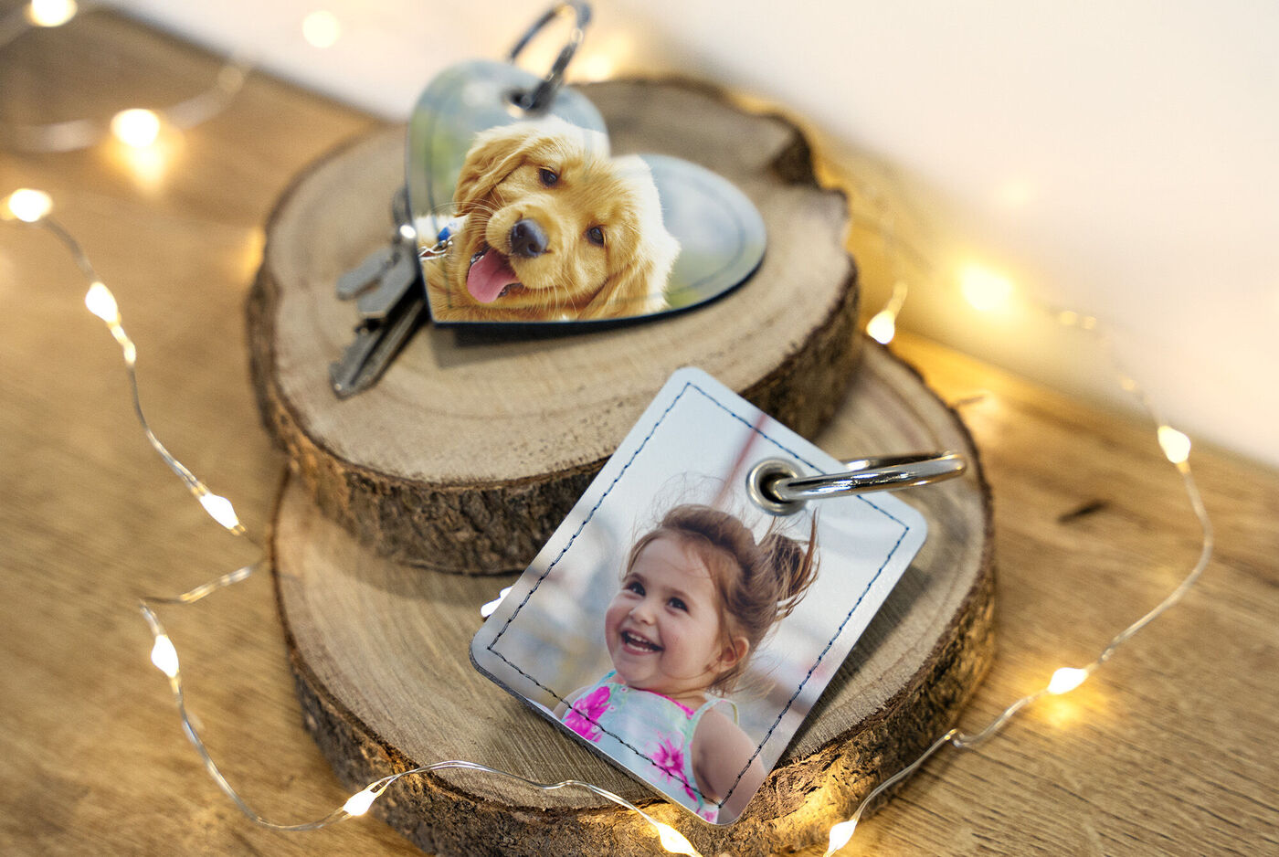 Deux porte-clés personnalisés sur un support en bois, l'un en forme de cœur avec une photo de chien et l'autre carré avec une photo de personne, entourés de lumières.