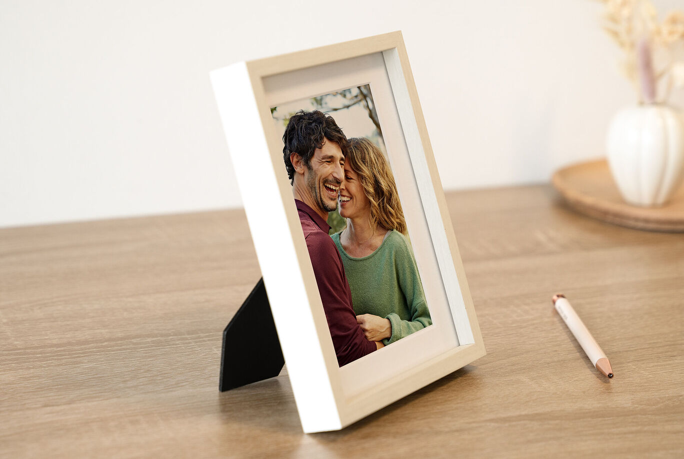 Une belle image dans un cadre blanc sur une table en bois, montrant un couple s'enlaçant. En arrière-plan, des couleurs douces et un vase de fleurs.