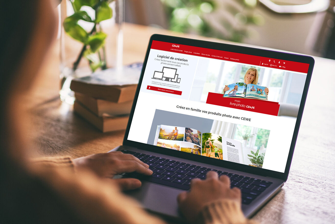 Une personne utilise un ordinateur portable sur une table avec des plantes et un album photo en arrière-plan.