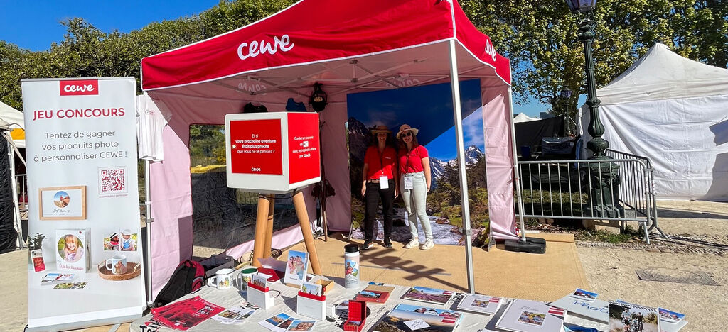 Un stand CEWE lors d'un événement avec des matériaux promotionnels, deux employées et une toile rouge.