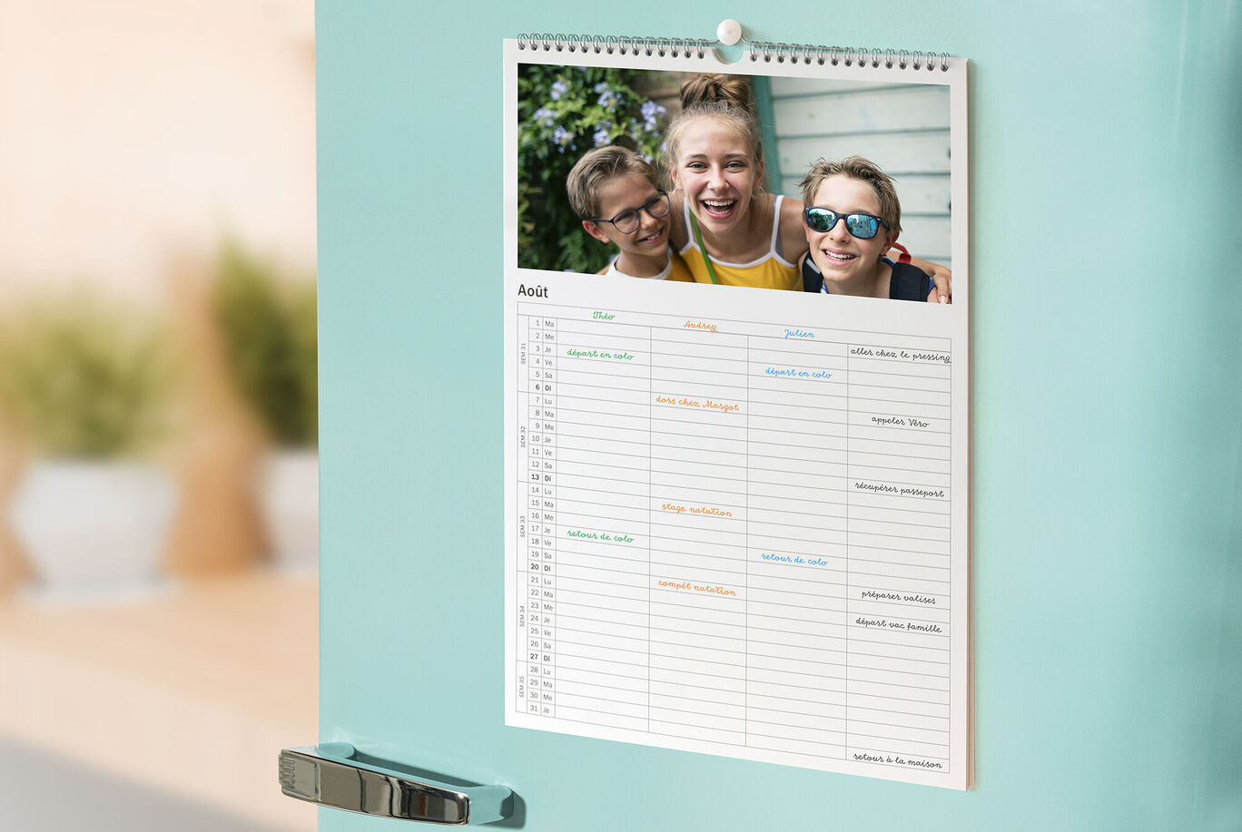 Un calendrier sur un frigo, avec un design coloré et une joyeuse photo de trois amis en arrière-plan.