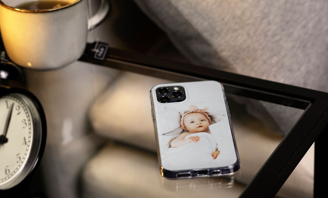 Un smartphone dans une coque de protection claire sur une table, entouré d'une horloge et d'une tasse.