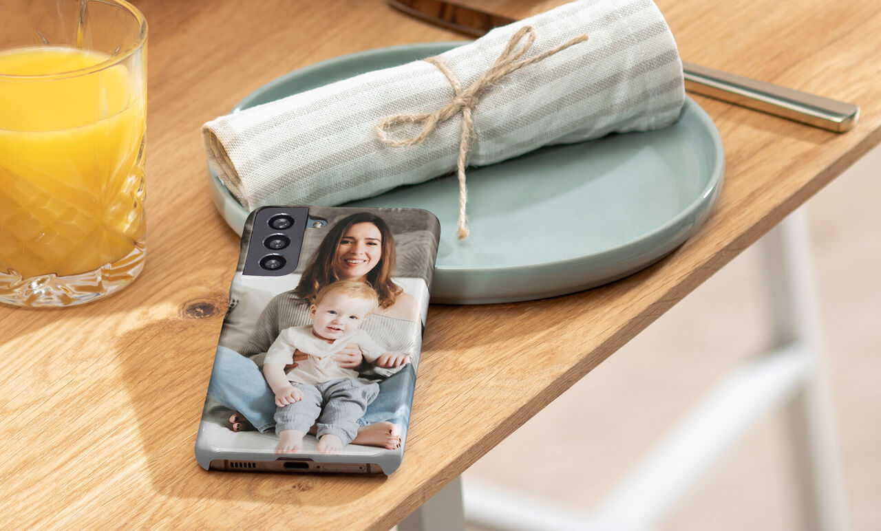 Un smartphone avec un design photo est posé sur une table, à côté d'un verre de jus d'orange et d'une assiette.